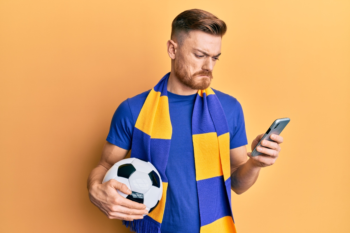 Anxious man holding a ball and checking betting results on a cellphone.